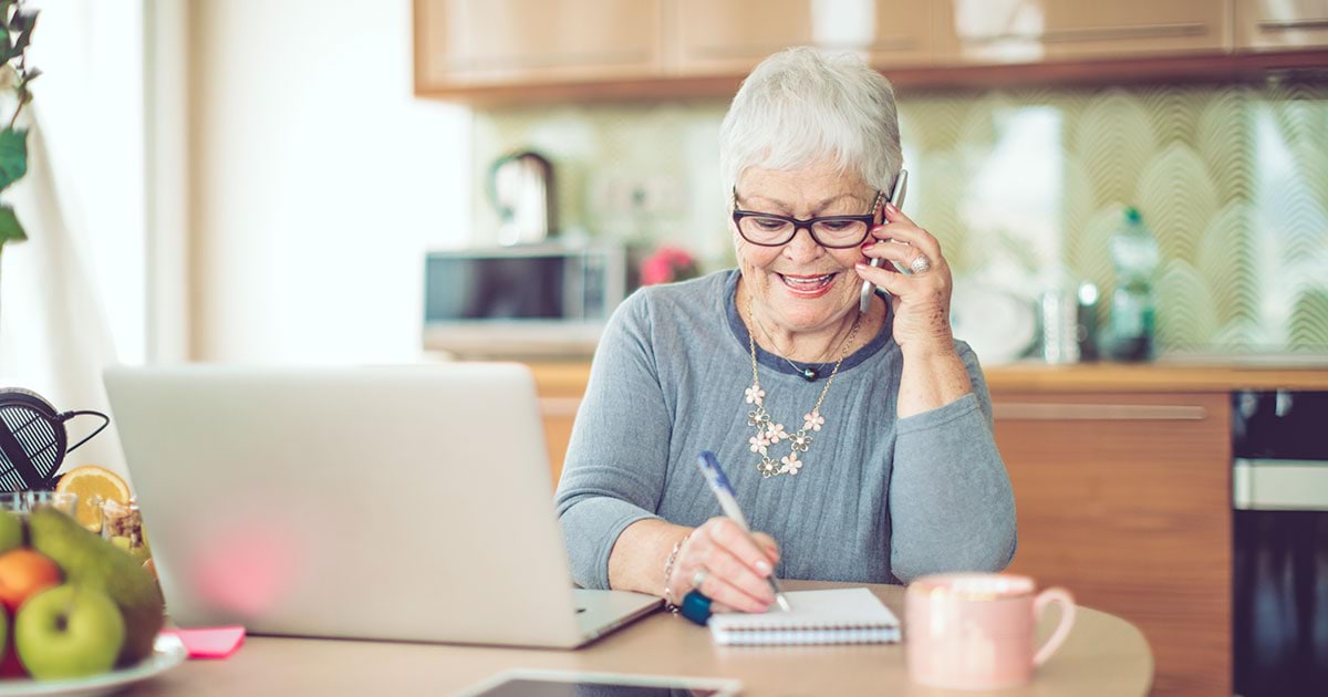 An older white woman with glasses takes notes next to her laptop at the table